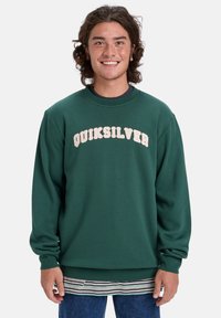 Zelený crewneck sveter s logom "QUIKSILVER" v odtieňoch krémovej, mäkká textúra, pruhované manžety a spodný lem, nosený cez pruhovanú košeľu.
