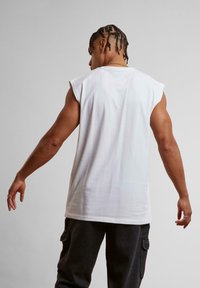 White sleeveless t-shirt with a relaxed fit, crew neckline, and smooth texture, worn with dark cargo pants. Subtle stitching detail at the back.
