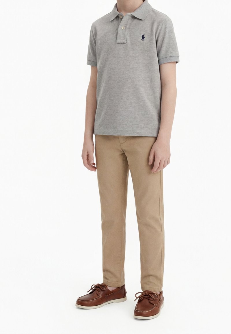Enfant debout portant un polo gris à manches courtes, un pantalon beige et des chaussures bateau marron sur fond blanc.