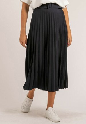 Femme portant une jupe midi plissée noire avec une ceinture assortie, des baskets blanches et une chemise blanche rentrée, debout sur un sol clair.