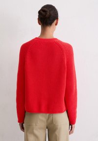 Czerwony sweter dzianinowy z okrągłym dekoltem, ryżowym splotem i raglanowymi rękawami. Charakteryzuje się skróconym krojem i jest zestawiony z jasnobeżowymi spodniami.