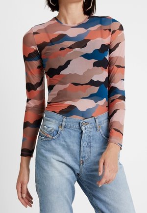 Kvinde iført en tætsiddende langærmet top med brun, orange, pink, blå og sort abstrakt mønster og lyseblå højtaljede jeans.