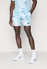 Heledad sinise värvi tie-dye lühikesed püksid sünteetilisest kangast, vasakul küljel väikese logoga, koos valgete sportlike kingade ja sokkidega.