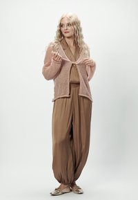 Jasnoróżowy, dziany kardigan z otwartym przodem, noszony na brązowym, jedwabnym kombinezonie. Całość dopełniają metaliczne buty.