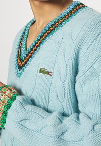 Lätt blått kabelstickat tröja med djup V-ringning och flerfärgad kant samt en liten grön Lacoste-logotyp på bröstet.