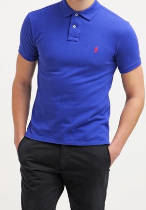 Homme portant un polo bleu ajusté avec un logo rouge sur la poitrine et un pantalon noir, les mains dans les poches, devant un fond uni.