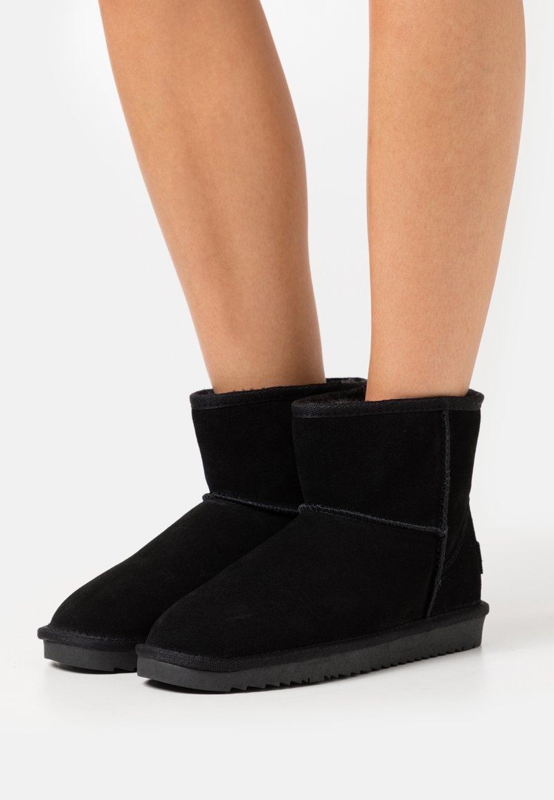 Stivali alla caviglia in suede nero con una morbida fodera interna, suola piatta in gomma e cuciture a contrasto. Design minimalista senza decorazioni.