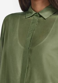 Chemise boutonnée semi-transparente couleur vert olive avec col, patte de boutonnage avant et léger éclat. Le tissu semble lisse avec une texture légère.