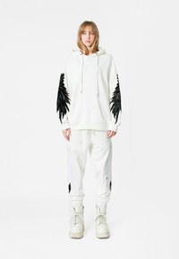 Sudadera blanca con capucha y adornos negros similares a plumas en las mangas, combinada con pantalones de chándal a juego que presentan diseños de alas plateadas. Botas blancas de plataforma.