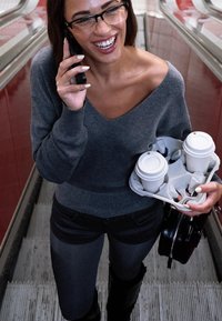 Sivi pulover, crne traperice, visoke crne čizme. Drži nosač za kavu s dva bijela šalica. Smiješi se, koristi pametni telefon dok je na pokretnim stepenicama.