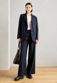 Blazer a righe navy e pantaloni a gamba larga, abbinati a una maglietta beige e scarpe con tacco nero. Gli accessori includono una borsa nera.