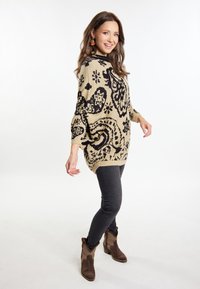 Maglione oversize beige con motivi neri intricati, abbinato a jeans scuri e stivaletti marroni. Tessuto morbido e vestibilità rilassata.