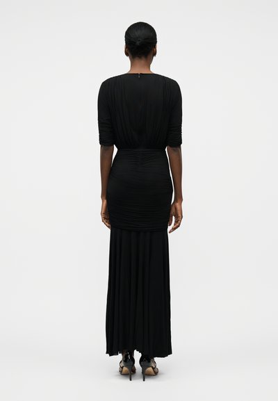 Ženska s temno poltjo in lasmi spetimi v figo, oblečena v dolgo črno nabrano obleko in črne sandale z visoko peto, stoji obrnjen hrbtno na belem ozadju.