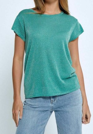 Tannengrünes Kurzarmoberteil aus strukturiertem, glitzerndem Stoff. Rundhalsausschnitt und lockere Passform, kombiniert mit hellblauen Jeans.