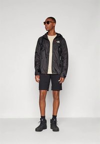 Mand iført solbriller, sort vindjakke, beige skjorte, sorte shorts, vandrestøvler og rygsæk, der står foran en hvid baggrund.