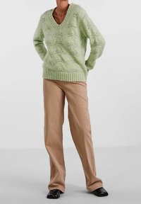 Pull vert clair en maille avec un col en V, présentant un motif texturé. Associé à un pantalon beige à jambes droites et des chaussures noires.