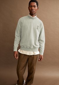 Lichtgroene sweatshirt met een hoge hals en een sleutelgatlogo, gelaagd over een wit shirt, gecombineerd met bruine cargobroek.