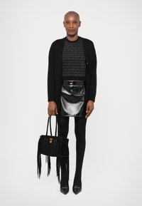 Schwarzer, gekürzter Pullover mit strukturiertem Muster, metallisch schwarzer Rock und schwarze Tasche mit Fransen. Schwarze Strumpfhosen und spitze Schuhe runden das Outfit ab.