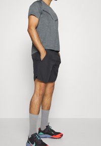 Mężczyzna w szarej, wzorzystej koszulce sportowej, czarnych szortach z kieszeniami, szarych skarpetkach do kostek i czarnych butach biegowych z czerwonymi i turkusowymi akcentami.