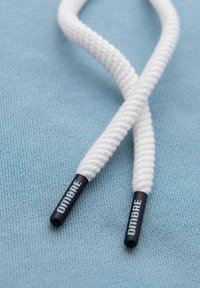 Zbliżenie na białe końcówki sznurowadeł z czarnymi agletami oznaczonymi "DIMBRE" na jasnoniebieskim tle z tkaniny.