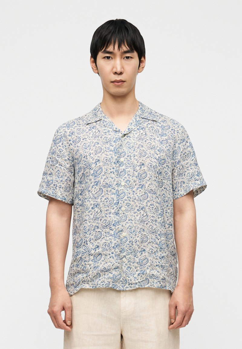 Chemise à manches courtes avec boutons, en tissu léger, présentant un motif floral bleu paisley sur fond beige, dotée d'un col camp et d'une coupe décontractée.