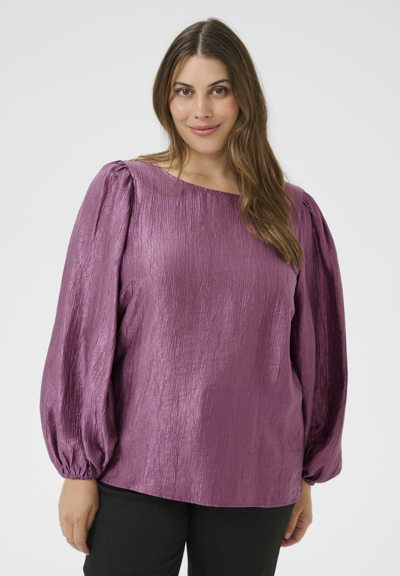 Donna con lunghi capelli castani che indossa una blusa viola lucida a maniche lunghe con maniche a palloncino e pantaloni neri, sorridendo leggermente, su uno sfondo bianco.