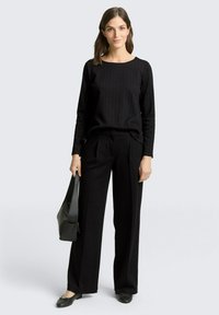 Czarna bluzka z długim rękawem i subtelnymi prążkami, zestawiona z czarnymi spodniami z wysokim stanem. Modelka trzyma czarną skórzaną torebkę.