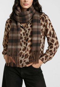 Donna che indossa una blusa con stampa leopardata marrone e beige, pantaloni neri e una sciarpa spessa a quadri marrone avvolta attorno al collo.