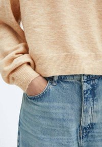 Leicht pfirsichfarbener Strickpullover mit umgeschlagenen Bündchen, kombiniert mit hochgeschnittenen, verwaschenen blauen Jeans aus Denim mit einer Vorderasche und Knopfverschluss.