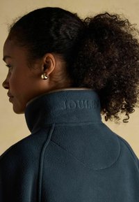 Žena s kučeravými vlasmi v copu nosí strieborné kruhové náušnice a tmavomodrú fleecovú bundu s vysokým golierom, na ktorom je vytlačené "JOULES."