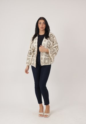 Mujer luciendo una chaqueta de crochet beige con estampado sobre un top negro y jeans skinny oscuros, con zapatos de tacón blancos abiertos en la punta, posando frente a un fondo liso.