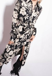 Kobieta w czarnej, kwiatowej sukience z długim rękawem i paskiem w talii, w czarnych butach na obcasie z frędzlami, pozująca na białym tle.