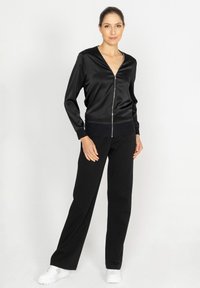 Czarna satynowa kurtka zapinana na zamek z dekoltem w serek i prążkowanymi mankietami w połączeniu z czarnymi spodniami o szerokich nogawkach i białymi trampkami na jednolitym tle.