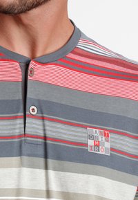 Camisa de manga corta a rayas en gris, rojo y beige con cuello tipo henley, dos botones y un parche con logotipo que presenta texto en rojo y gris.