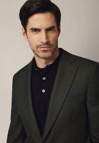 Ciemnozielona marynarka o teksturowanym wykończeniu i wyciętymi klapami, noszona na czarnej koszulce polo z białymi guzikami.