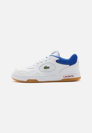 Weißer Sneaker mit blauer Kragen, Gummisohle, Lacoste-Logo an der Seite und "LACOSTE"-Text an der Mittelsohle vor neutralem Hintergrund.
