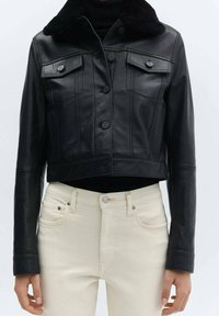 Chaqueta corta de cuero negra con cierre de botón y dos bolsillos en el pecho. Cuenta con un cuello forrado de piel y una textura suave. Combinada con jeans de color claro.