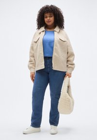 Bēšas krāsas vilnas jaka ar apkakli un ielaidnēm, kopā ar zilu garroku kreklu, ziliem džinsiem un krēmkrāsas somu. Balti sporta apavi.