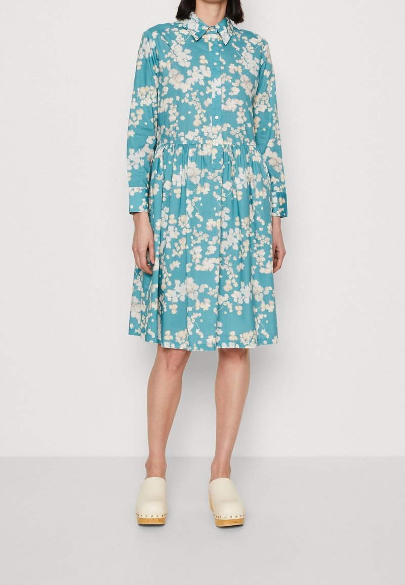 Turkos blommig klänning med långa ärmar, åtsittande överdel och plisserad kjol. Har en krage och knäppning med knappar. Bärs med krämfärgade träskor.