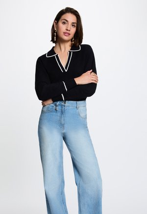 Vrouw met kort bruin haar die een zwart topje met lange mouwen en witte bies draagt en lichtblauwe high-waisted jeans, staand met gekruiste armen.