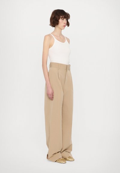 Beige, hoch taillierte, weite Hose mit einem Gürtel in der Taille, kombiniert mit einem weißen Tanktop und goldenen, geflochtenen Slippern. Glatte Stofftextur.