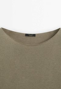 Haut vert olive avec un col large et arrondi, en matière tricotée texturée. Étiquette visible près du col, indiquant la marque et l'origine.