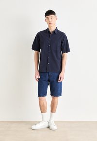 Marineblå kortærmet skjorte med knapper og en lomme, parret med denim-shorts og hvide sneakers, samt ribbede hvide sokker.