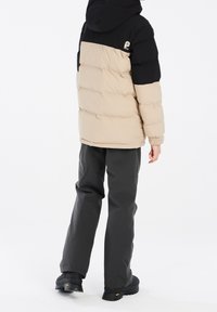 Doudoune avec des sections beiges et noires, dotée d'une capuche, de poignets élastiques et d'un logo. Associée à un pantalon gris foncé et des bottes noires.