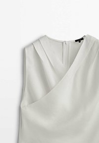 Blouse blanche sans manches avec col en V et devant drapé façon portefeuille, dotée d'une fermeture éclair au dos et d'un détail plissé aux épaules.