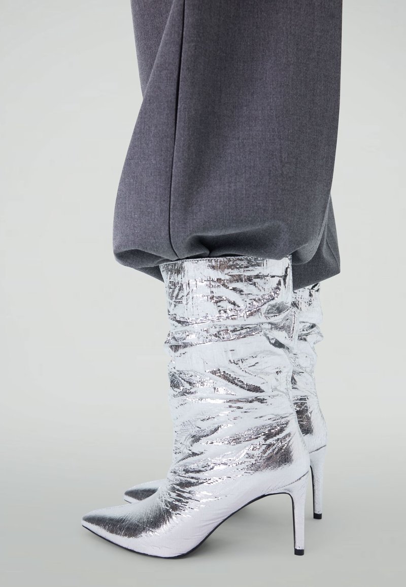 Bottes argentées montant jusqu'au genou avec une texture métallique froissée et un bout pointu. Les talons sont fins, créant une silhouette élégante.