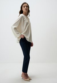 Blouse crème à col rond et manches longues, associée à un pantalon tapered bleu marine foncé et des chaussures à talons nus. Texture de tissu lisse.
