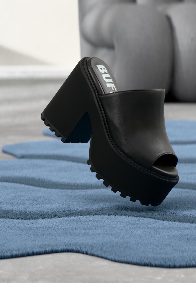 Mule plateforme noire à bout ouvert avec semelle épaisse à crampons reposant sur un tapis bleu texturé à l'intérieur, près d'un fauteuil gris.