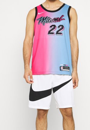 Mann trägt ein Miami Heat Basketballtrikot in Pink und Blau mit der Nummer 22 und weiße Nike-Shorts mit großen schwarzen Swooshes.