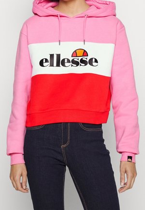 Kobieta ubrana w kolorową bluzę ellesse z kapturem w odcieniach różu, bieli i czerwieni z sznurkami, w połączeniu z ciemnoniebieskimi dżinsami.